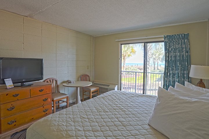 A hotel room with a bed, TV, table, and chair in front of a sliding glass door leading to a balcony.