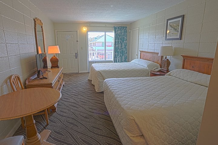 A hotel room with two double beds, a wooden table and chairs, and a window with a view of another building.