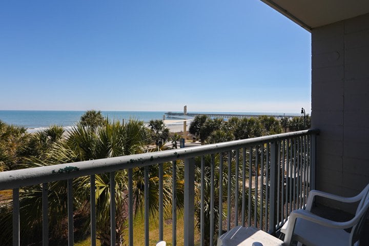 A balcony with a metal railing overlooking the ocean and beach with palm trees.